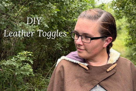 A woman wears an Overland Cloak that is fastened by two large handmade leather toggles, rather than buttons. She is standing on a wooded path. A title reads DIY Leather Toggles.