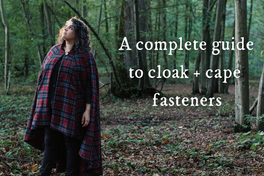A woman wears a red and blue plaid Cascades Cloak in the middle of a forest; text reads A Complete Guide To Cloak and Cape Fasteners.